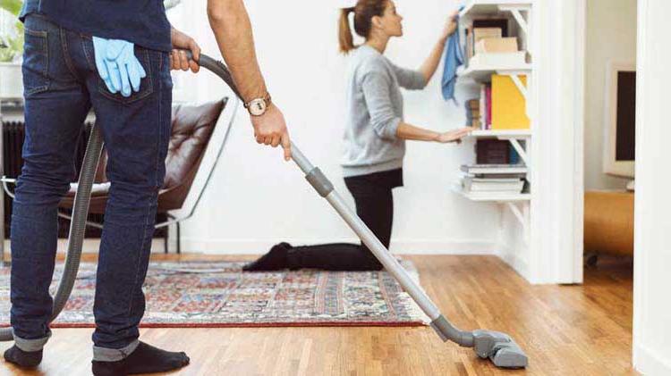 Woman dusting a bookshelf and man vacuuming to get rid of dust in the home.